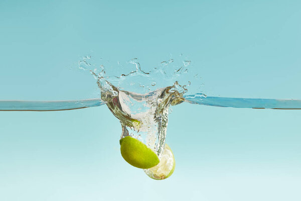 спелые половинки лайма падают глубоко в воду с брызгами на голубом фоне
