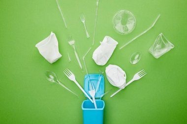 yeşil arka plan üzerinde mavi geri dönüşüm kutusu yakınında tek kullanımlık plastik bardak, çatal ve kaşık üst görünümü