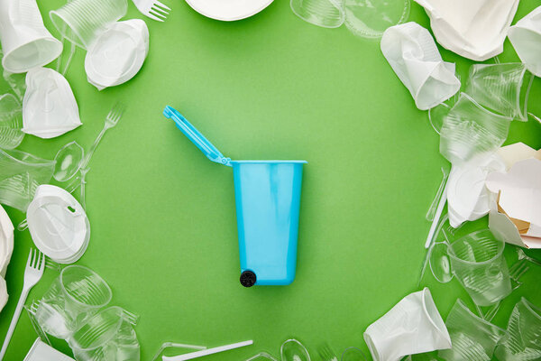 top view of blue recycle bin between crumpled plastic cups, forks, plates and cardboard container on green