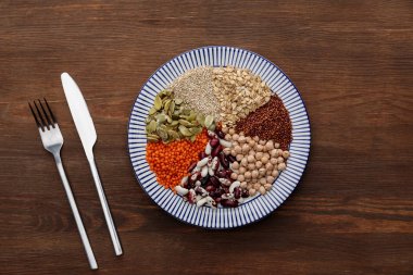 ahşap yüzeyde çiğ mercimek, nohut, kinoa, yulaf ezmesi, fasulye ve kabak çekirdeği ile çizgili plaka yakın çatal ve bıçak üst görünümü