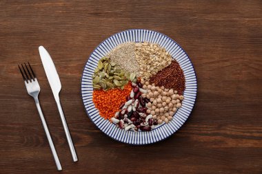 top view of fork and knife near striped plate with raw lentil, chickpea, quinoa, oatmeal, beans and pumpkin seeds on wooden surface clipart