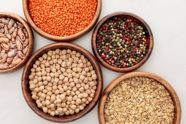 top view of wooden bowls with chickpea, oatmeal, red lentil, beans and peppercorns on white marble surface clipart