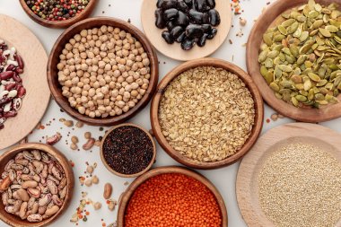 top view of wooden bowls with diverse beans, oatmeal, red lentil, peppercorns, pumpkin seeds and chickpea on white marble surface with scattered grains clipart