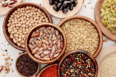 top view of wooden bowls with diverse beans, peppercorns, pumpkin seeds, oatmeal, quinoa and chickpea on white marble surface with scattered grains clipart