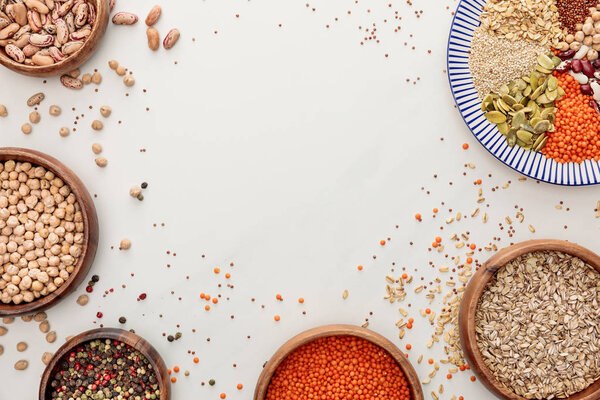 top view of bowls and plate with raw lentil, quinoa, oatmeal, beans, peppercorns and pumpkin seeds on marble surface with scattered grains and copy space