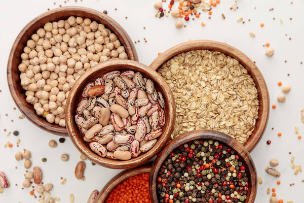 top view of wooden bowls with oatmeal, red lentil, beans, peppercorns and chickpea on white marble surface with scattered beans