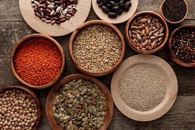 top view of bowls with raw oatmeal, red lentil, various beans, quinoa, chickpea, peppercorns and pumpkin seeds on wooden surface clipart