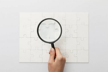 cropped view of woman holding magnifying glass under jigsaw puzzle with lost piece on white background clipart