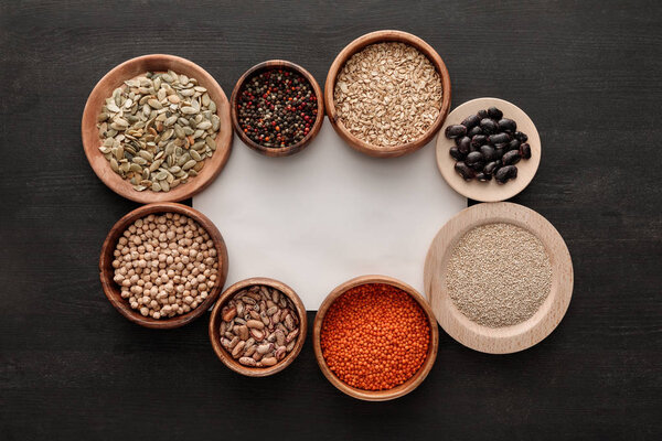 верхний вид чистой белой бумаги рядом с деревянными тарелками и чашами с бобами, злаками, пряностями и тыквенными семечками на темном деревянном столе
