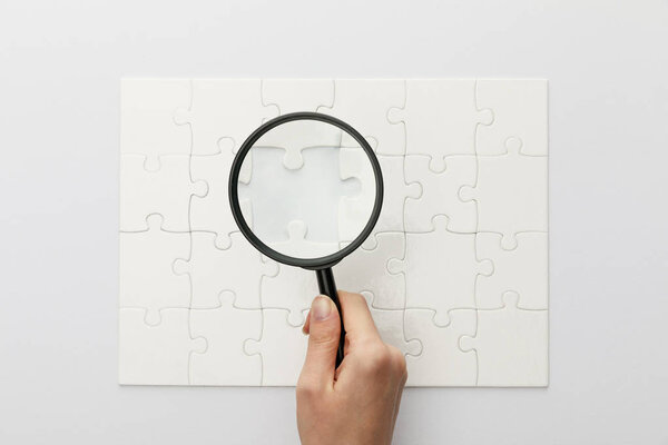 cropped view of woman holding magnifying glass under jigsaw puzzle with lost piece on white background