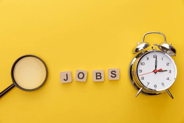 top view of magnifying glass, clock and cardboard squares with jobs inscription on yellow background