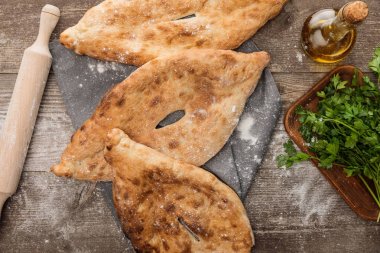lavaş ekmeğinin oklavanın yanında gri havlu üzerine üst görünümü, dağınık unlu tahta masa üzerine taze maydanoz ve zeytinyağı