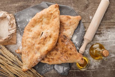 lavaş ekmeğinin tahta masa üzerinde oklava, un paketi, sivri ve zeytinyağının yanında gri havlu üzerinde üst görünümü