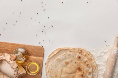 top view of flat lavash bread near wooden cutting board with flour and olive oil on white surface with peppercorns clipart