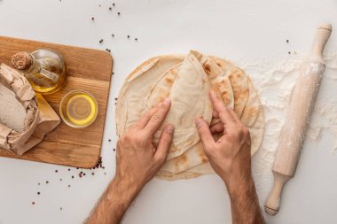 cropped view of man wrapping flat lavash bread near rolling pin and wooden cutting board with flour and olive oil on white surface with peppercorns clipart