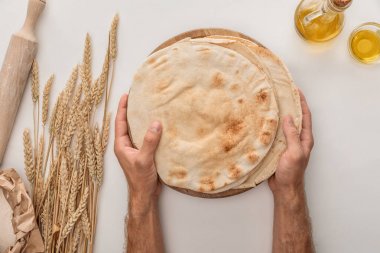 cropped view of man holding flat lavash bread near wheat spikes, rolling pin and olive oil on white surface clipart