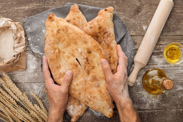 cropped view of man holding lavash bread on gray towel near rolling pin, flour package, wheat spikes and olive oil on wooden table