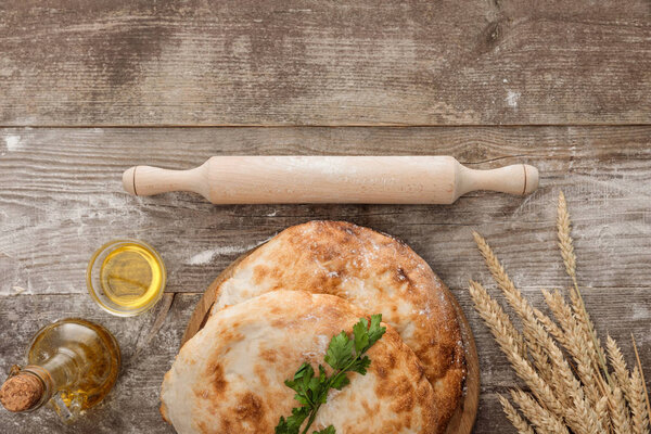 top view of lavash bread near wheat spikes, rolling pin, fresh parsley and bottle of olive oil on wooden table