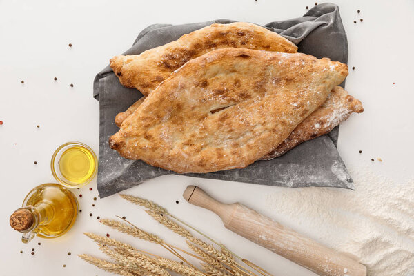 top view of lavash bread on gray towel near wheat spikes, rolling pin and olive oil on white surface with peppercorns