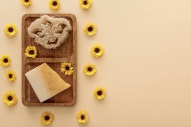 çiçeklerle bej arka plan üzerinde loofah ve sabun parçası ile kahverengi ahşap sabunluk üst görünümü