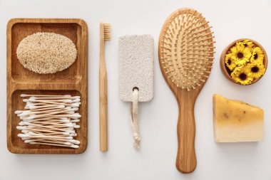 flat lay with wooden dish with cotton swabs and loofah near toothbrush, hairbrush, piece of soap, pumice stone and cup with flowers on white background clipart
