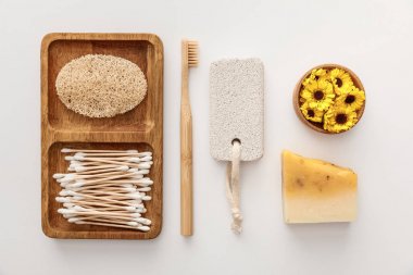 flat lay with wooden dish with cotton swabs and loofah near toothbrush, piece of soap, pumice stone and cup of flowers on white background clipart