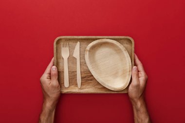 cropped view of man holding natural rectangular wooden dish with plate, fork and knife on red background clipart