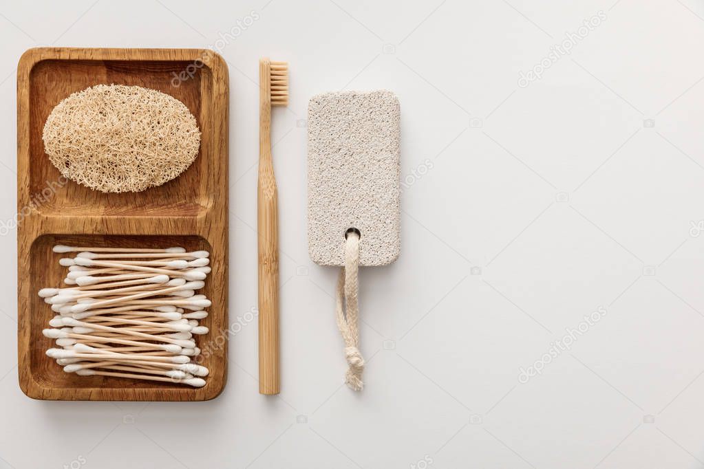 Flat lay with wooden dish with cotton swabs and loofah near toothbrush and pumice stone on white background