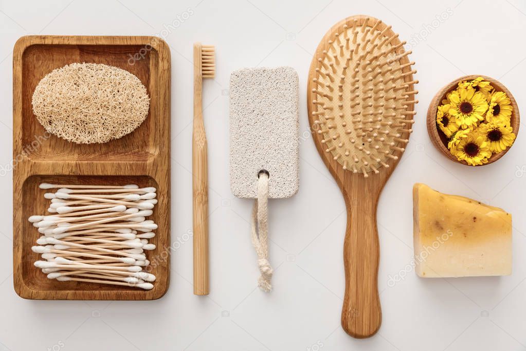 Flat lay with wooden dish with cotton swabs and loofah near toothbrush, hairbrush, piece of soap, pumice stone and cup with flowers on white background