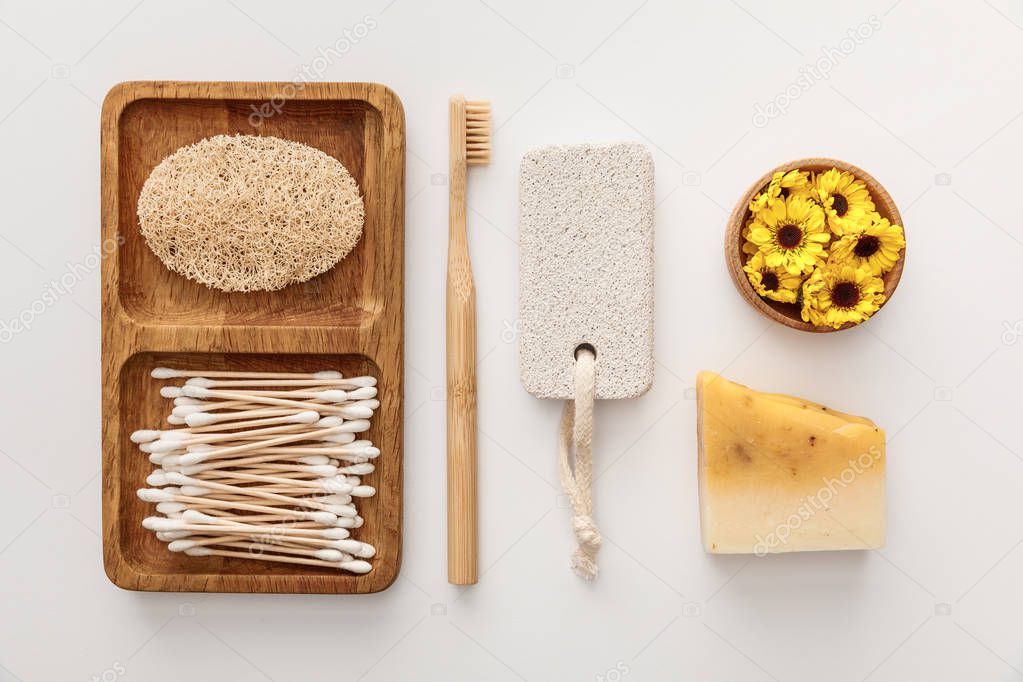 Flat lay with wooden dish with cotton swabs and loofah near toothbrush, piece of soap, pumice stone and cup of flowers on white background