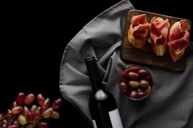 Beyaz etiketli şarap şişesinin üst görüntüsü olgunlaşmış üzüm, zeytin ve dilimlenmiş prosciutto yanında siyah üzerine izole edilmiş ekmek.