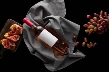 top view of bottle of rose with blank white label on grey napkin near ripe grape and sliced prosciutto on baguette isolated on black clipart