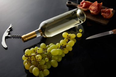 bottle with white wine near grape, knife, corkscrew and sliced prosciutto on baguette on black background clipart