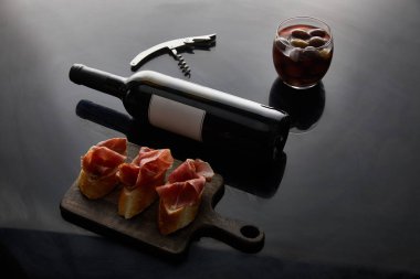 wine bottle with blank label near sliced prosciutto on baguette, corkscrew and olives on black background clipart