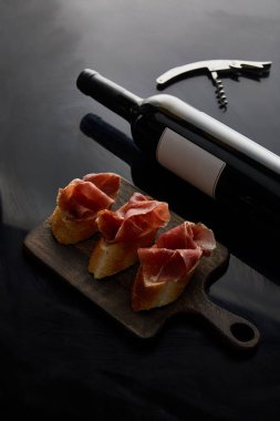wine bottle with blank label near sliced prosciutto on baguette, corkscrew on black background clipart
