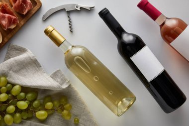 Üzüm, tirbuşon ve beyaz arka planda dilimlenmiş jambon yanında beyaz, kırmızı ve gül şarabı olan şişelerin üst görüntüsü.