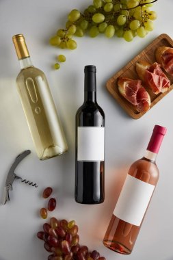Üzüm, tirbuşon ve beyaz arka planda dilimlenmiş jambon yanında beyaz, kırmızı ve gül şarabı olan şişelerin üst görüntüsü.