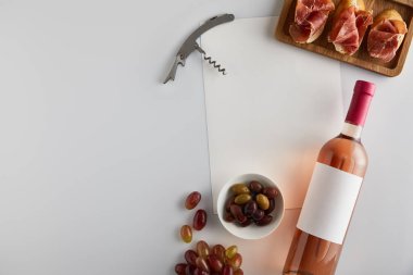 Üzüm, tirbuşon, zeytin, boş kağıt ve beyaz arka planda dilimlenmiş ekmek üzerine doğranmış jambon olan şişe manzarası.