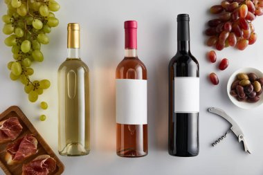 Üzüm, tirbuşon, zeytin ve beyaz arka planda dilimlenmiş jambon yanında beyaz, gül ve kırmızı şarap bulunan şişelerin üst görüntüsü.