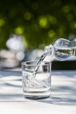 fresh clean water pouring from bottle in transparent glass at sunny day on wooden table clipart