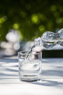 fresh clean water pouring from bottle in transparent glass at sunny day on wooden table clipart