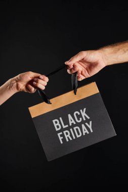 cropped view of woman and man holding paper shopping bag with black Friday lettering isolated on black clipart