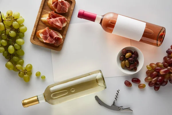 top view of bottles with white and rose wine near grape, corkscrew, blank paper, olives and sliced prosciutto on baguette on white background