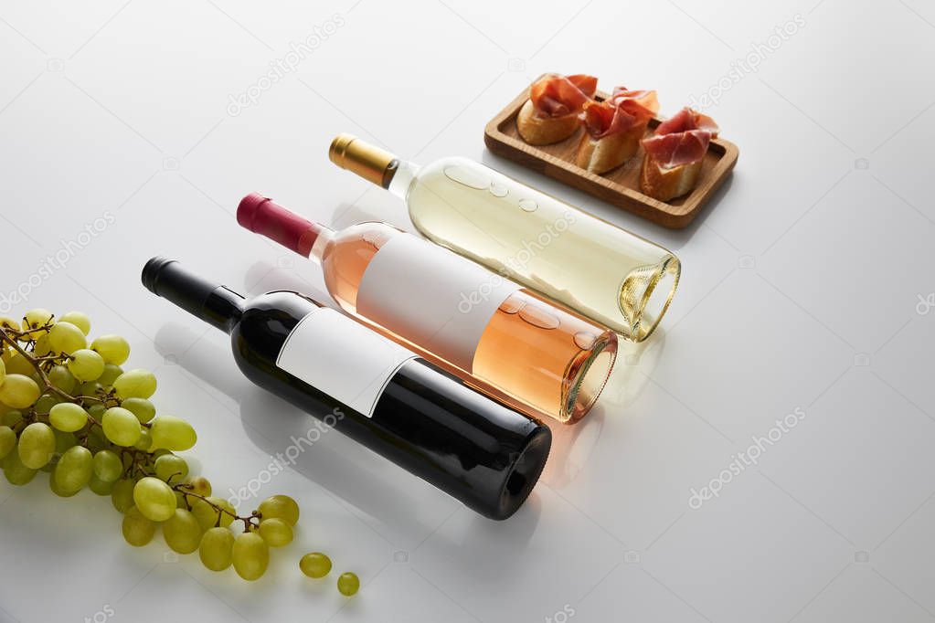 bottles with white, rose and red wine near grape and sliced prosciutto on baguette on white background
