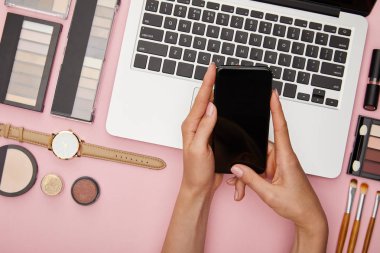 Bilgisayarın yanında boş ekranlı akıllı telefonu olan ve pembe üzerine izole edilmiş dekoratif kozmetik ürünleri olan bir kadının üst görüntüsü. 