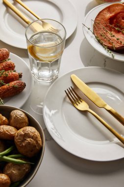 Fırında sebzeli beyaz tabaklar, altın çatallar ve bıçaklar, beyaz mermer masada limonlu su bardağı.