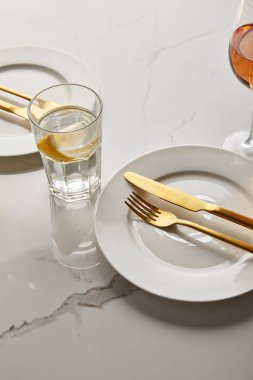 Beyaz tabakların yanında limon suyu, altın çatallar ve mermer masada bıçaklar.