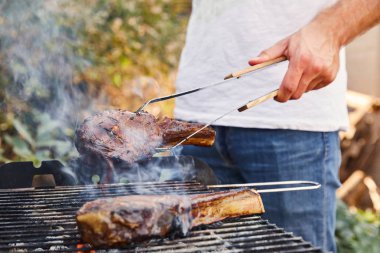 cropped view of man with tweezers grilling meat on barbecue grid  clipart