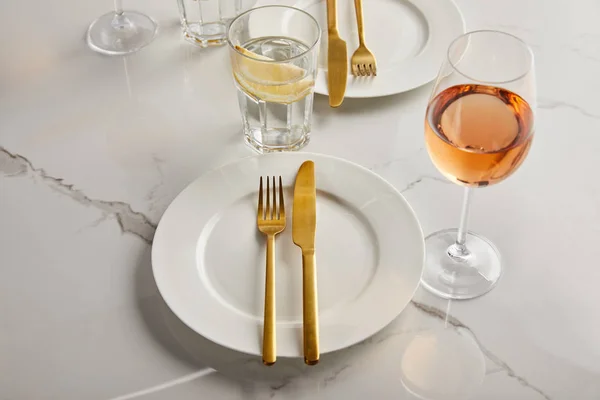 大理石のテーブルの上にローズワインとレモンの水とガラスの近くの白いプレート上の黄金のナイフとフォーク — ストック写真