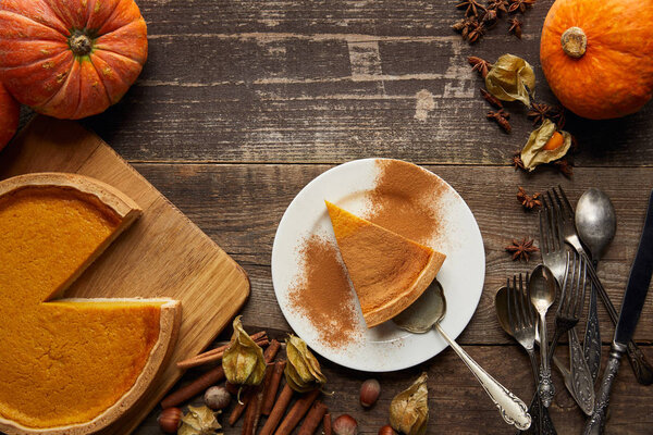 top view of tasty pumpkin pie near whole pumpkins, cutlery, cinnamon, hazelnuts and physalis on dark wooden surface